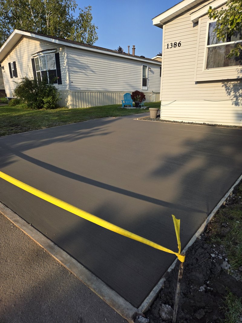 Dark broom-finished driveway slab beside a white mobile home